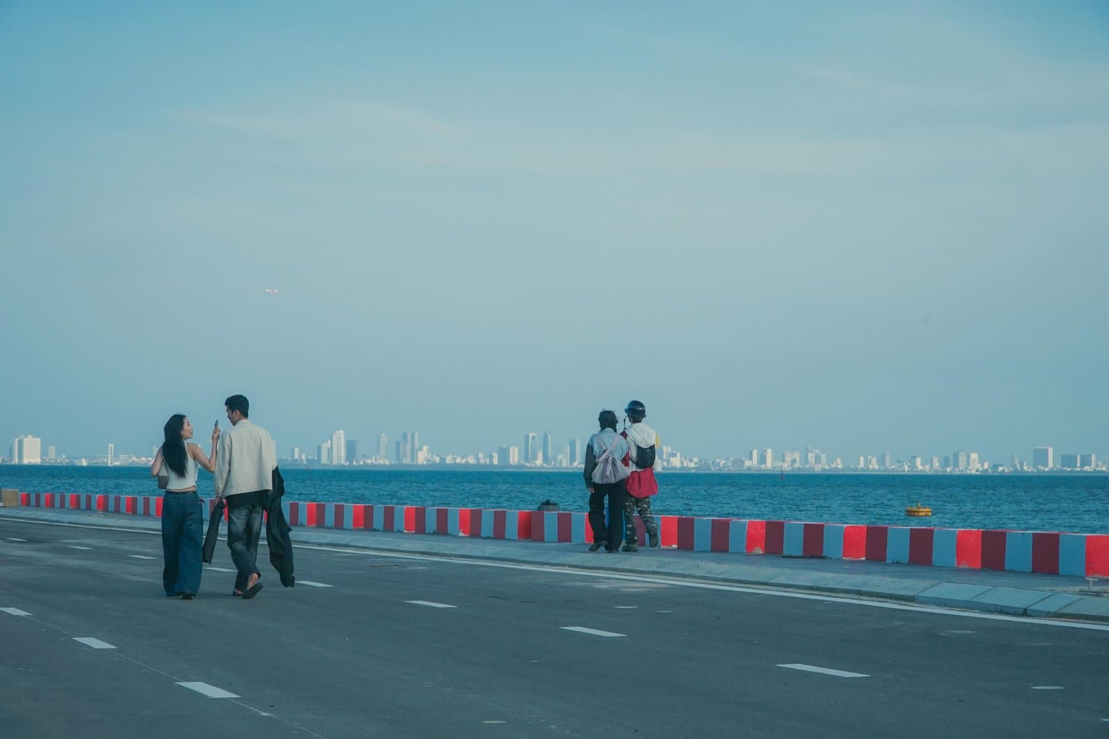 Two couples enjoy a stroll on a seaside promenade in Da Nang, Vietnam, with the city skyline in the distance.